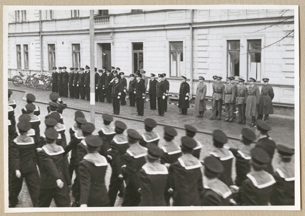 Uniformsklädda flottister marscherar på Drottninggatan medan militärer av högre rang, från marinen och armén, står uppställda utanför bataljon Sparres kanslibyggnad.