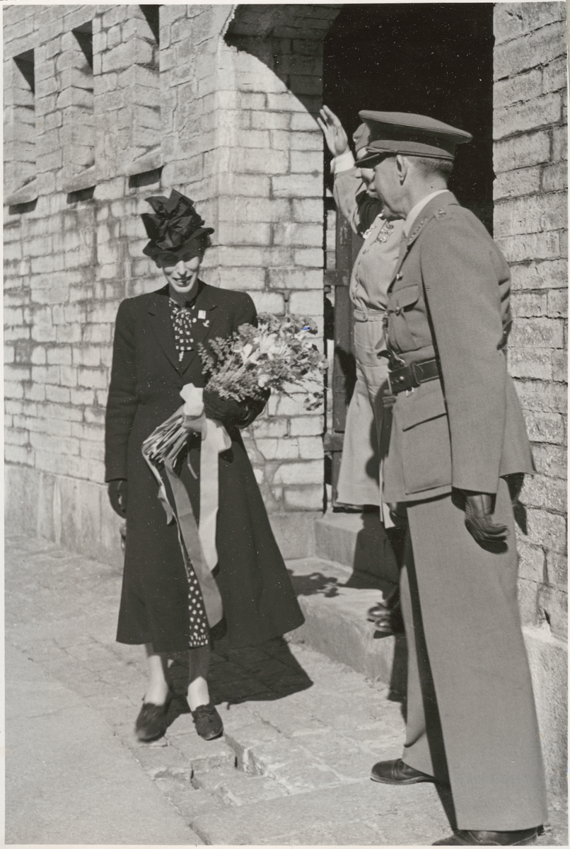 Drottning Louise, klädd i mörk dräkt och hatt samt med en blombukett i famnen, står i rundhamnen tillhörande Kungsholms fort tillsammans med en lotta och en militär från armén, båda klädda i uniform.