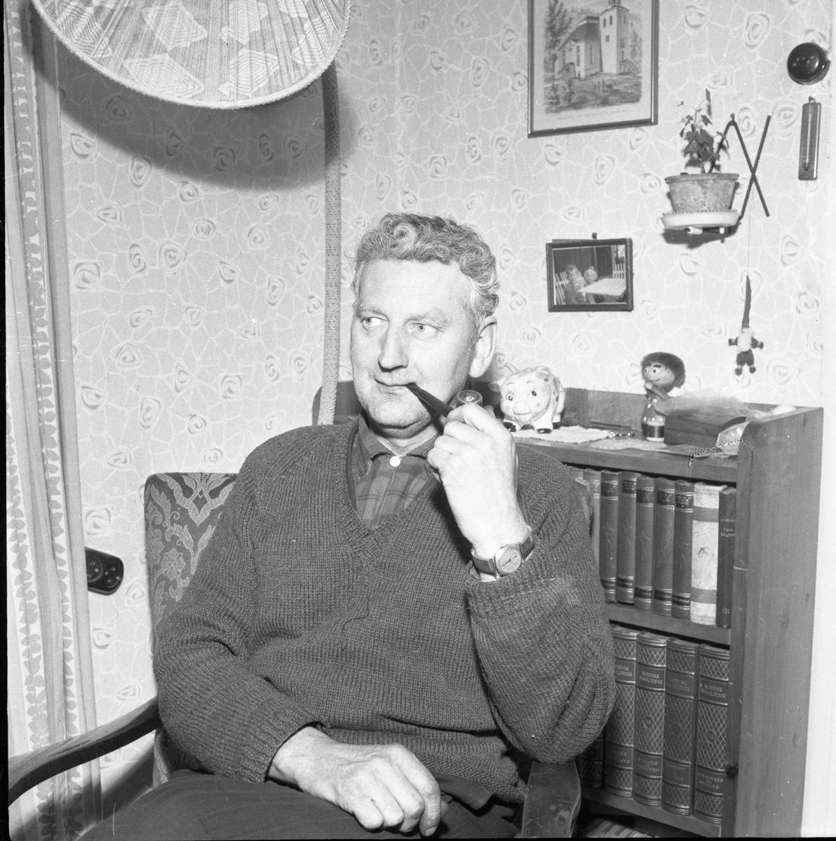 Skorstensfejarmästare Rune Skoglund sitter in en fåtölj med pipa i munnen. Bakom honom en låg bokhylla med bl a Nordisk Familjebok.