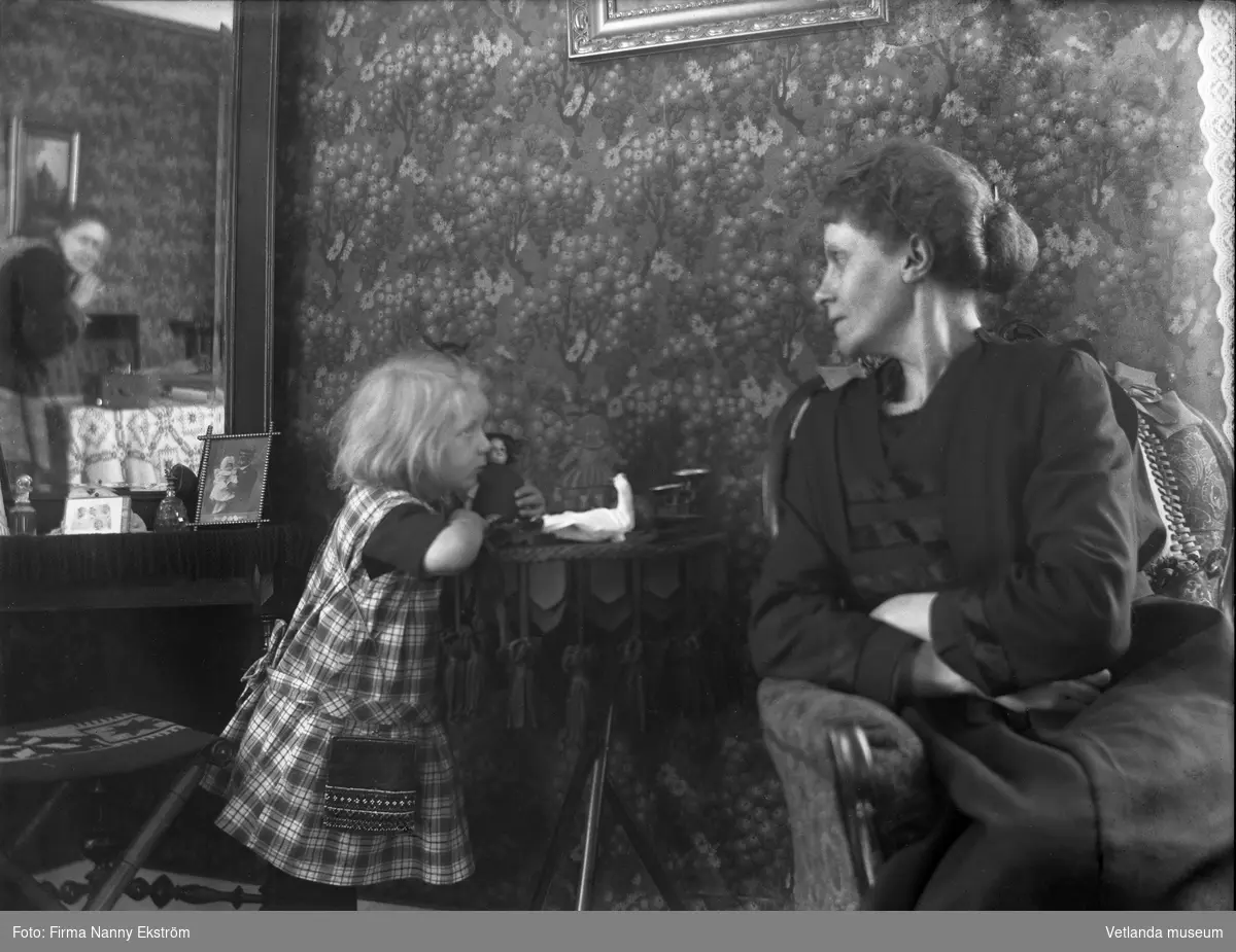 Kvinna och barn med docka.
Fotografen Nanny Ekström kan skymtas i spegeln med sin kamera.