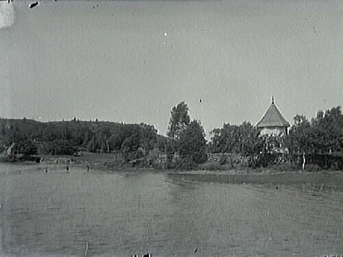 Vy över sjön mot Nösslinges muromgärdade kyrka i Halland. Endast tornet syns och lite av kyrkan till höger i bild (på brukskopian).