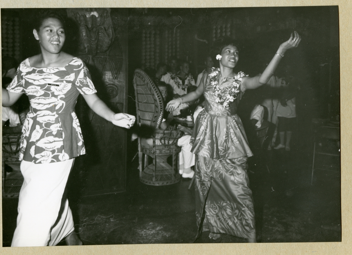 Bilden föreställer två kvinnor som framför en dans. I bakgrunden syns uniformsklädda män som åskådare. Bilden är tagen i Samoa i samband med minfartyget Älvsnabbens långresa 1966-1967.