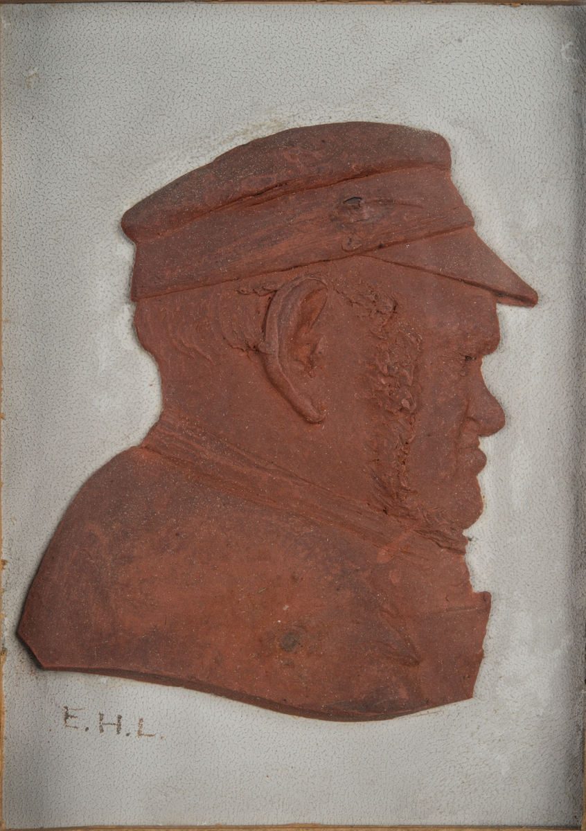 Relief i brun plastelin av "Gubben C. S. Ihrfors född 1832, död 1901", under glas, i svart ram.