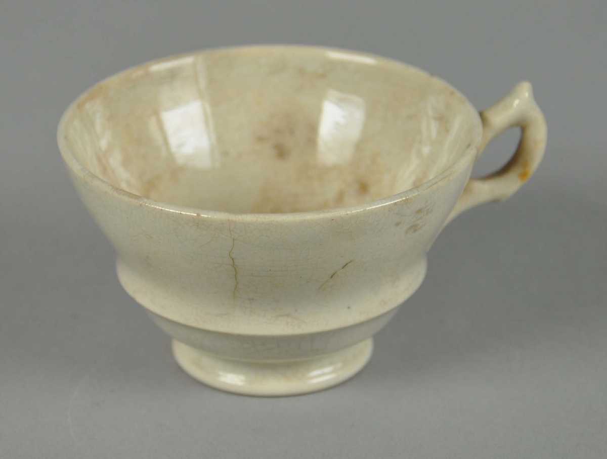 Kopp av glassert keramikk. Koppen er hvit og har hank med dekorelementer. Rundt korpus går det en utstående, profilert linje.