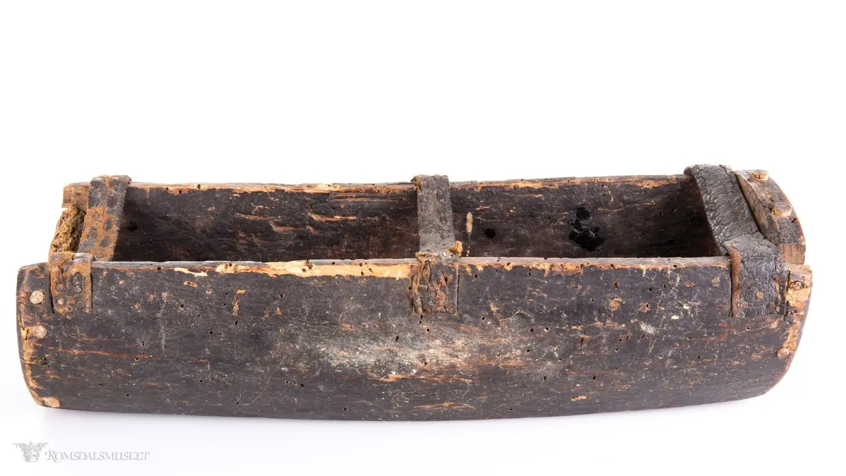 Uthulet trestokk som er høvlet flat i bunnen og har påsatte endestykker som er festet med trenagler. Tre lærremmer er spikret fast på tvers av åpningen. Rester av bek i bunnen.