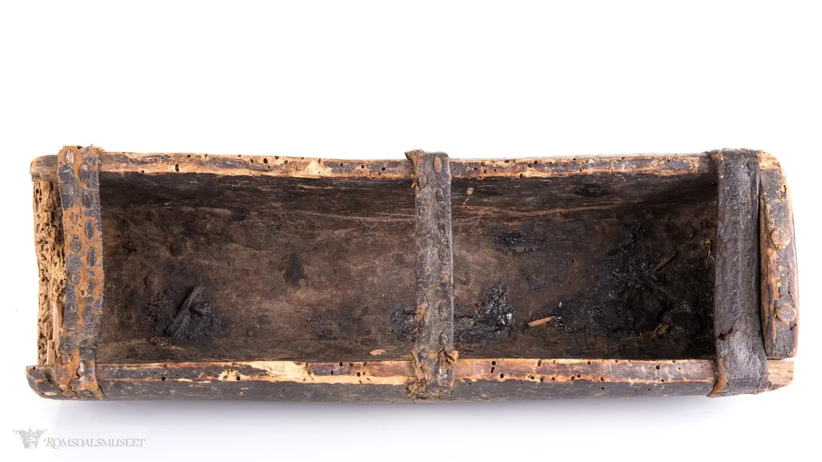 Uthulet trestokk som er høvlet flat i bunnen og har påsatte endestykker som er festet med trenagler. Tre lærremmer er spikret fast på tvers av åpningen. Rester av bek i bunnen.