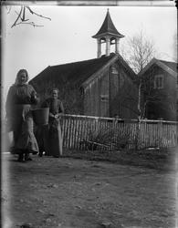 To uidentifiserte kvinner med et stabbur i bakgrunnen.