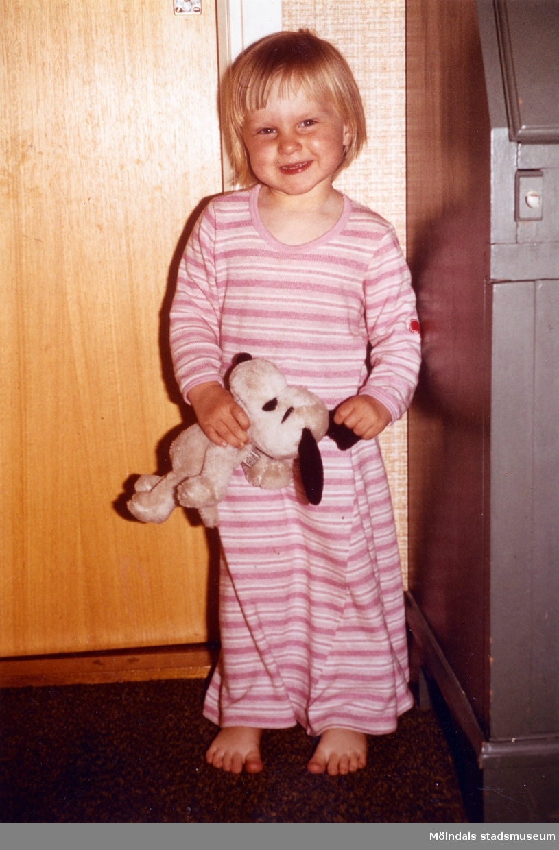 Hon är klädd i ett vitt- och rosarandigt nattlinne och står framför en innevägg. I händerna håller Karin ett gosedjur (Snobben). Troligtvis början av 1980-talet.