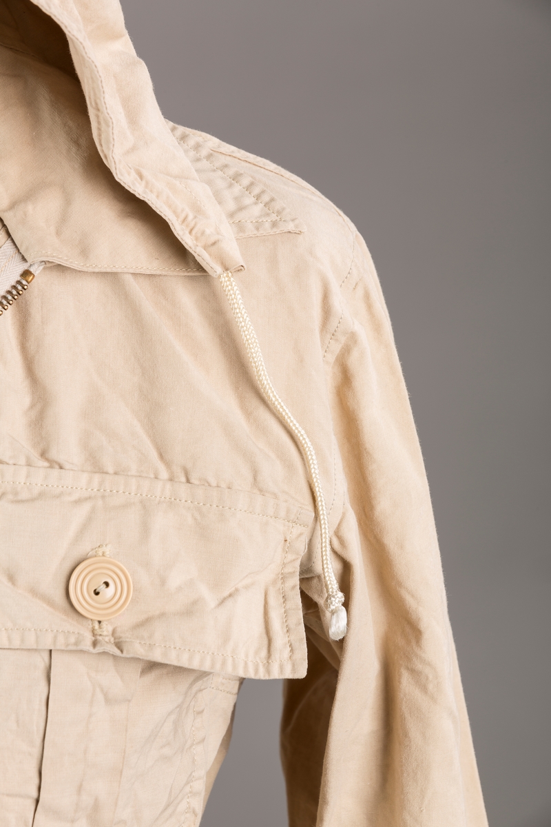 Form: Vanlig anorakkfasong med strikk i livet. 2 brystlommer med knapp.
Hette med glidelås på topp midt, snøring.
Glidelås helt ned foran.
Innermansjett med strikk i ermene.