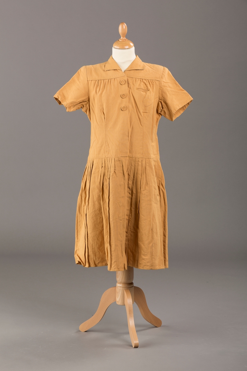 Form: Korte ermer, livet rynket til bærestykke foran, plissert skjørt, tre pynteknapper, en trykknapp, forlomme.