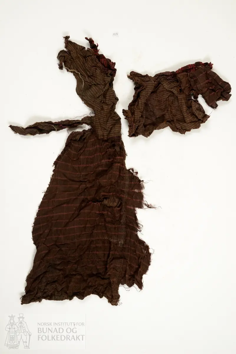 Svært utslitt del av ein stakk med linning, nederkant og ei metallhekte i eine enden av linning. Nedanfor denne delen av linning er det sauma fald som truleg er splitten midt framme på stakken. Av tre ulike verkensstoff, lerretsbinding med bomull i renning, ull i innslag. Stripete stoff i brun og lilla, svært utydeleg lillafargar. Fleire små hol etter mus, har vore nytta som stappefille.