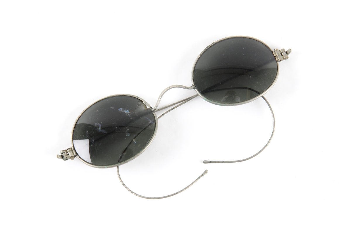 Solbrille med ovale og mørk grå glass. Tynn metallinnfatning med hengsler og to stenger.