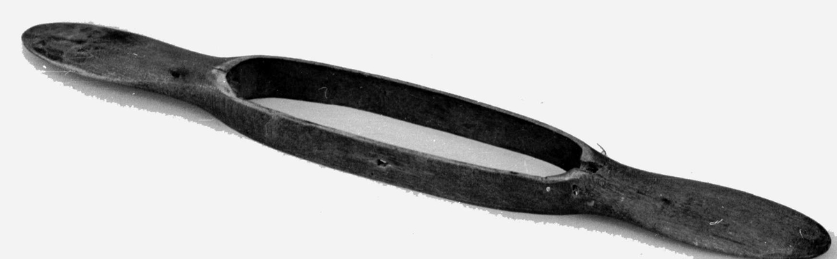 Vevskyttel av tre med lange håndtak på hver side. I midten er det skåret ut et hull, med plass til trådspolen.