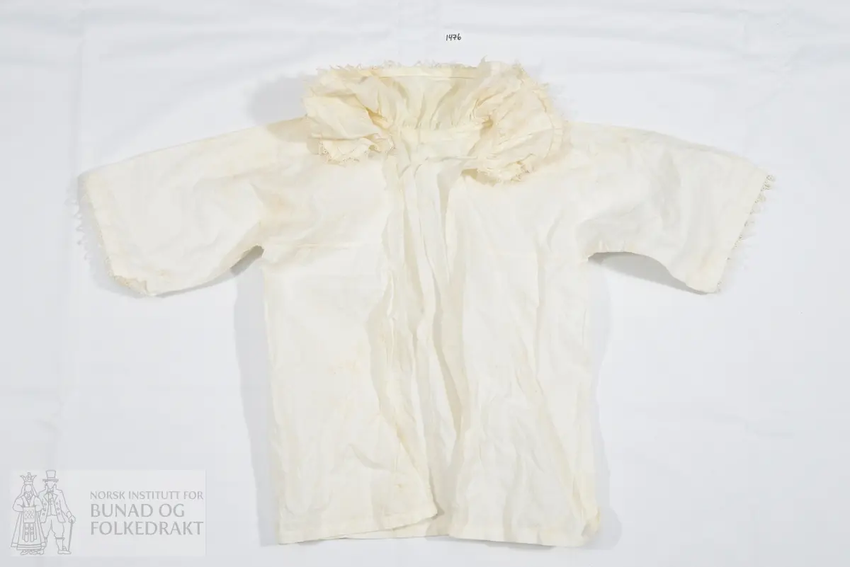 Barneskjorte. Hvitt bomullslerret. Krage med nuperellekant og hullfald er rynket til bolen i små rynker. Løpegang med knytebånd i halsen. Åpen hele veien ned midt foran. Lukkes med knyting i halsen. Ermene er rette og vide, er ikke isydd, men henger fast i ryggen og et bærestykke foran. Denne delen er skåret i ett og sydd til et stykke foran, hvor sømmen er en fortsettelse av sømmen under ermet. Ermene er kantet med nupereller. Kun maskinsøm.