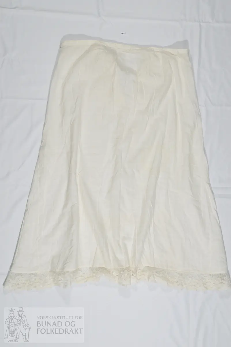 Understakk i hvit bomull. Kantet nederst med kjøpeblonde som trolig også er av bomull. Understakken har en smal linning øverst og lukkes med knytting. Sydd sammen av to høyder stoff som er lagt i små rynker til linningen. Ved linningen finner vi også to små innsnitt.  Understakken har i tillegg 2 isydde kiler fra omtrent knehøyde, disse er noe ujevne og har ikke helt samme høyde. Linningen har en skjøt midt bak. Maskinsøm