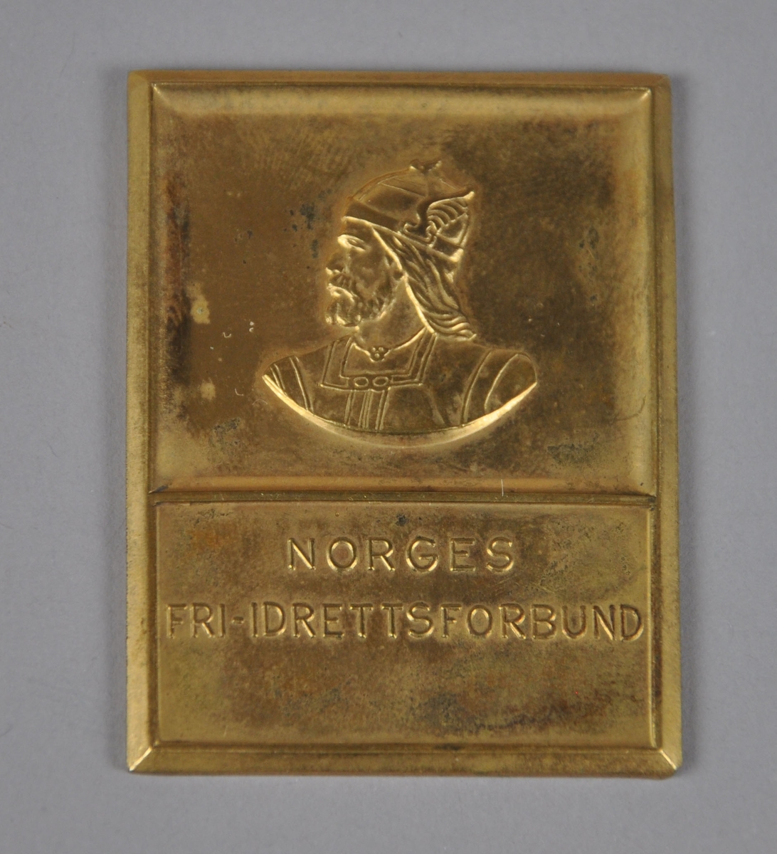 Medalje / plakett med bilde av en viking med hjelm og inskripsjon.
Plaketten ligger i eske med gjenstandsnr. OLM-08236