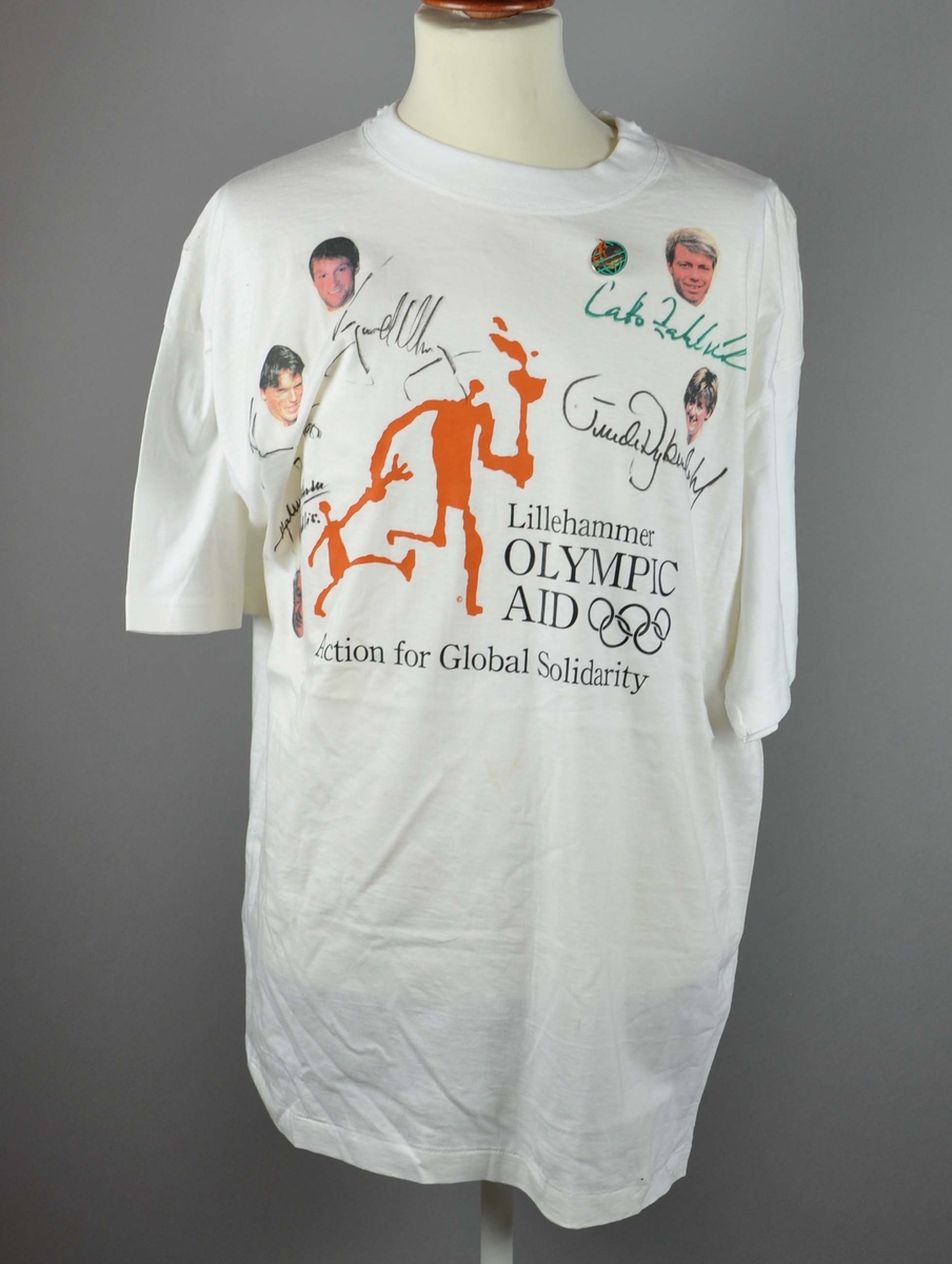 T-skjorte med logo for Lillehammer Olympic Aid og signaturer og bilder av Vegard Ullvang, Cato Zahl Pedersen, Johan O. Koss, Trude Dybendahl og Hjalmar Anderssen "Hjallis". Det er festet en nål/merke/pins for Olympic Aid på t-skjorten