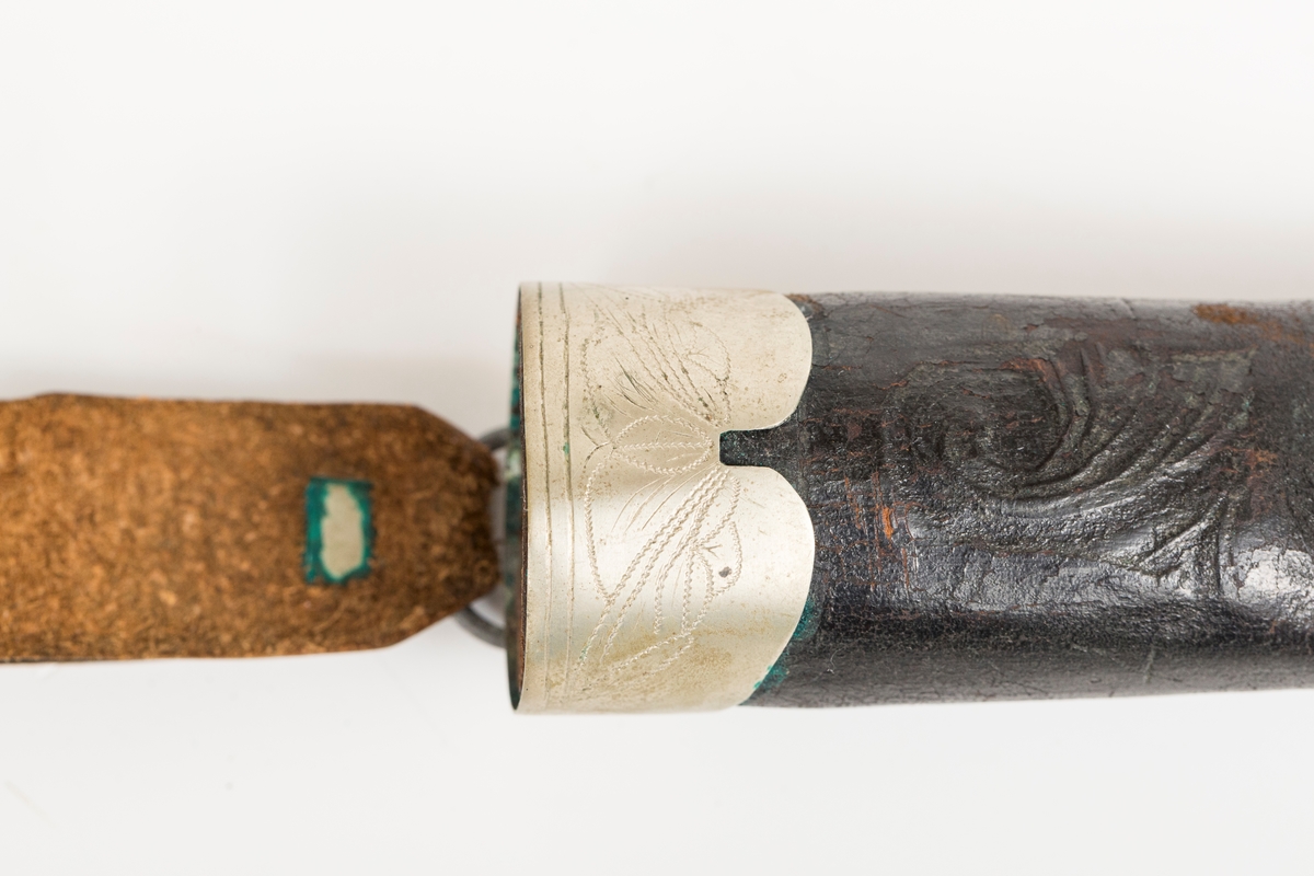 Håndsydd lærslire med holker oppe og nede. Slire og stropp av separate lærstykker. Sliren har enkel  pautet dekor. Holkene er håndloddet nysølv (sansynligvis) med enkel trambulert dekor. Toppholken har påloddet hengsle og D-ring til feste for slirestropp.