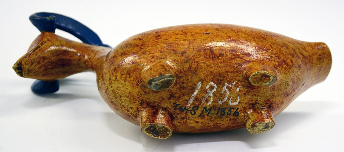 Kane. Fra protokollen: Kane, saak. "ølbøkaar", av træ. Kroppen nærmest mindende om gris + and, med 4 avstumpede ben og avstumpet hale. Hovedet bærer 2 foroverbøiede, fritliggende væderhorn (værhorn), som med sin forreste krumning hviler paa kjæven; hovedet forøvrigt væderlignende. Sen maling, kirketræsfarvet krop, blaa horn, hvit inside.