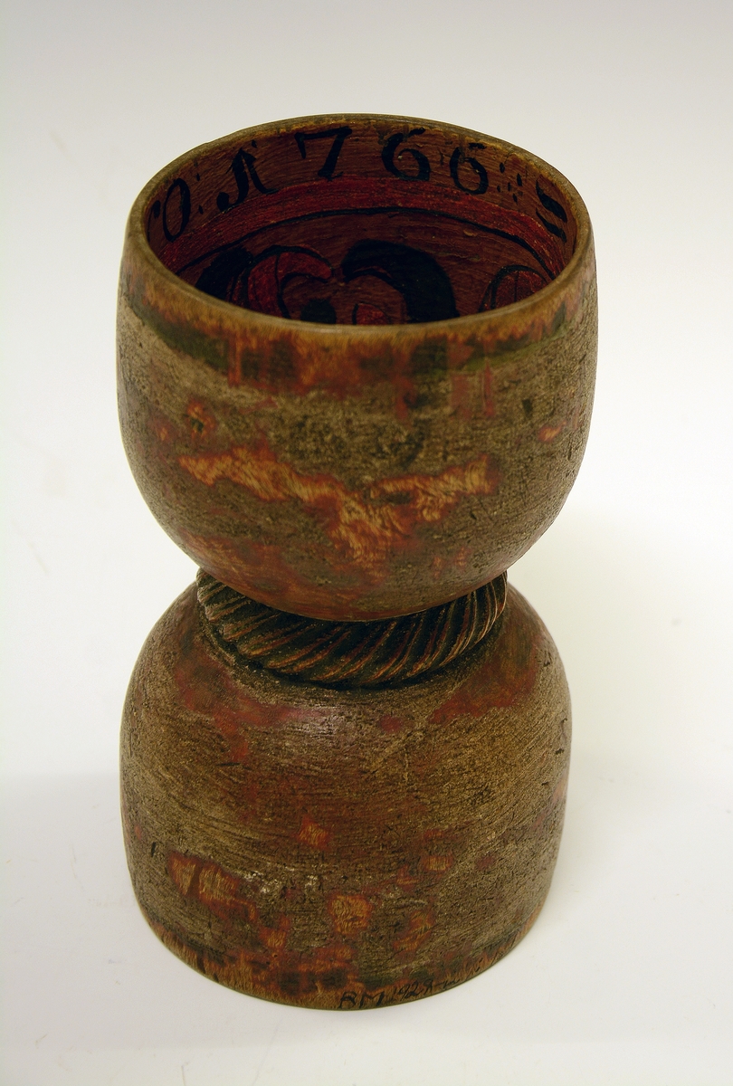 Dobbeltbeger. Fra protokollen: Bugge svarva av bjørk, maala. Baae staupi av paa lag same storleik, innsnørde med eit toglaga band. Utanpaa er den graa-raude maalingi noko avsliti, inni er rosemaala paa gul grunn med raude og grøne rosur, svart konturer. I det eine staupet er ein liten raud botnblom, og fraa denne gjeng mjuke rislur med tytebærblad paa. Kring tromi inni denne innskrift: "Bugen bruges i Fleng af Pige og Dreng". I det hitt staupet er maala eit stort firblad danna av 4 tulipanlaga blomar ut fraa ein botnblom. Millom tulipanane tytebærblad. Innskrift kring tromi inni "O.A.S.M. ANNO 1766"