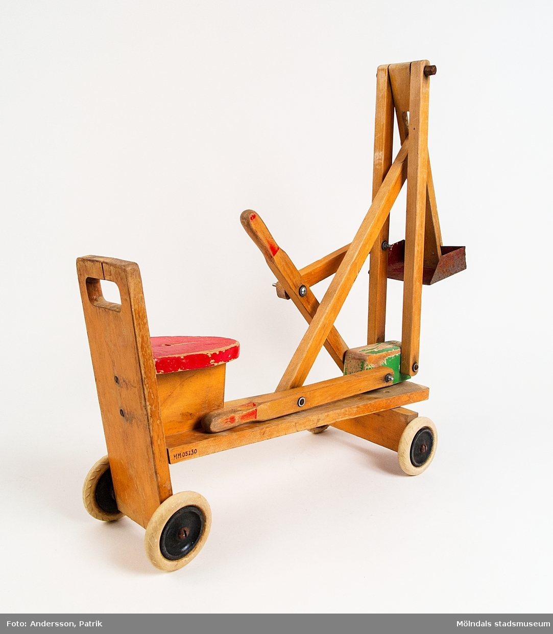 Grävskopa. Tillverkad av klarlackat trä med färglagda detaljer och röd sits. Fyra hjul med gummidäck. Grävskopan regleras av ett barn med hjälp av två spakar för lek i sandlåda.