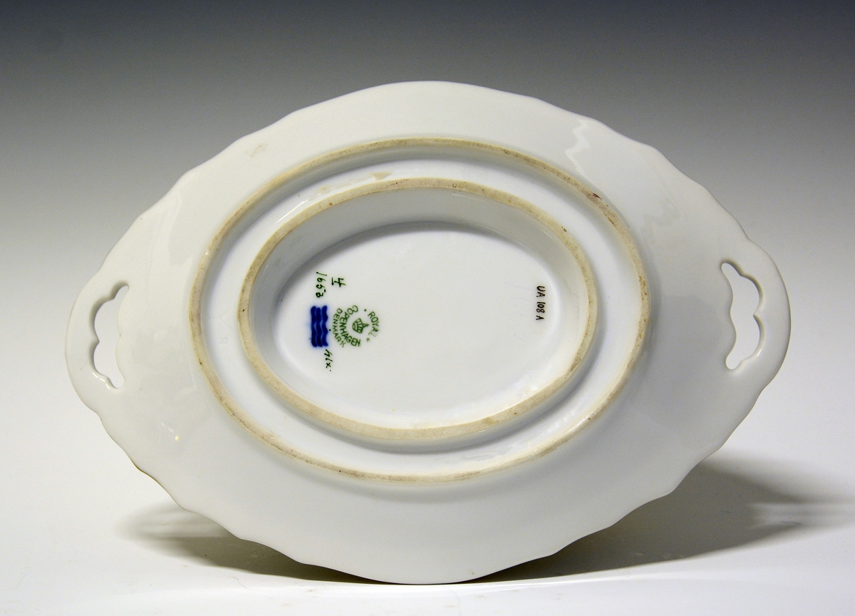 Sauseterrin av porselen dekrorert med håndmalt blomsterdekor og modellert figur av et barn med blåseintstrument som lokknapp. Hanker på hver siden med skjellform. Alle kanter er markert med gull.