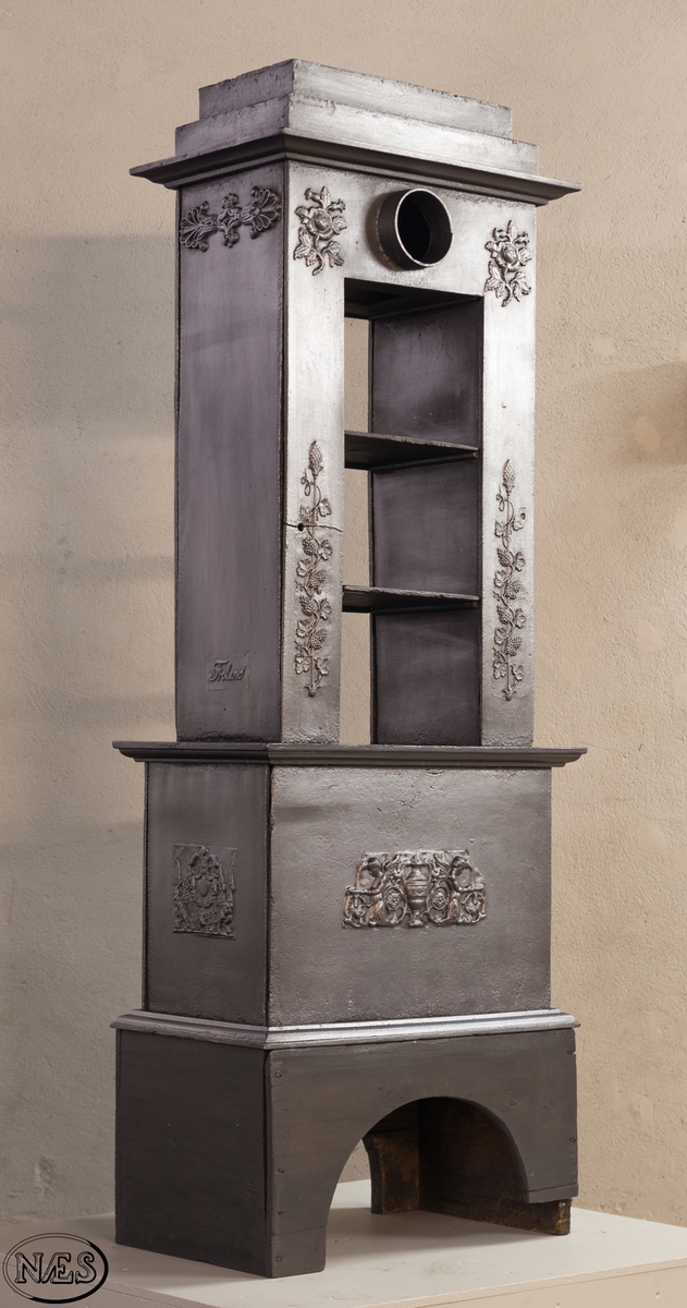 Ovnen består av to avdelinger. Underdelen har et frembuet midtparti med kamskjell flankert av sjødyr på hver side. På kortsidene skjold, løvehode og symmetriske ørner. På bakplate en krukke med symmetriske engler.
Overdelen er høy, firkantet med tredelt åpning. Empire ornamentikk og avtrappet topp. Øverst på frontplaten symetriske fabeldyr (pegasus) vendt mot hverandre.