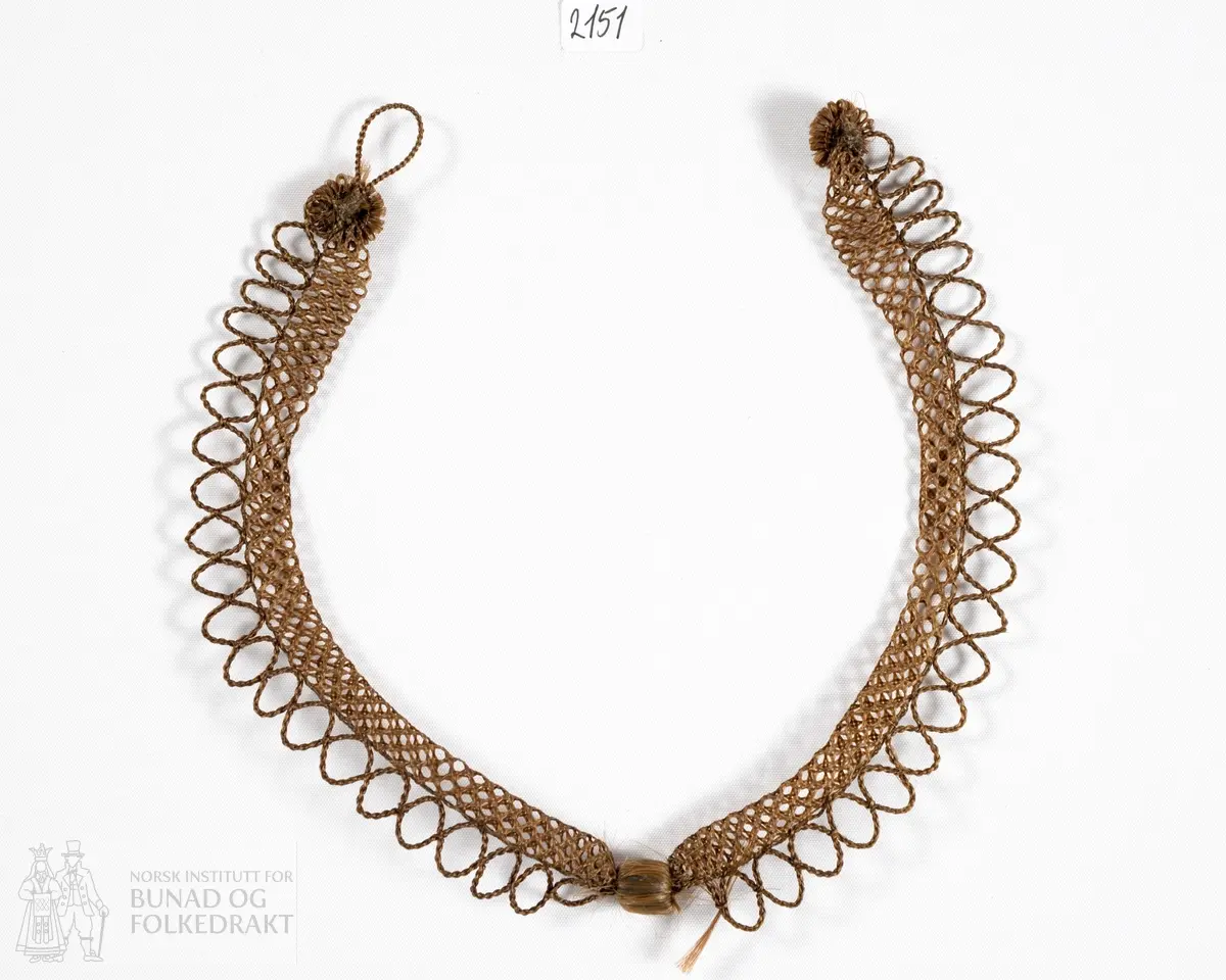 Smykke laget av menneskehår. En nettingformet klave kantet med løkker. Lukkes bak med en knapp og hempe som også er av hår.