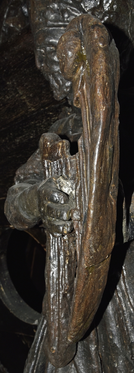 Skulptur utformad som en harpspelande man, med skägg och långt hår. Skägget är svagt krusigt och håret faller i lockar ned mot axlarna. Huvudet är vridet mot höger. Mannen står i knäande ställning, med höger ben något framskjutet och är iförd romersk rustning av typ muskelpansar - Thorax - med nedhängande lambrequinliknande och läderimiterande flikar ovanpå den tjocka livklädnaden eller tunican.

Figuren är iförd en stövelliknande fotbeklädnad med tjocka skaftprydnader. På huvudet förefaller han bära en slags mössa kring vilken en öppen krona trätts. I händerna, som hålls lyfta framför bröstet, håller han ett litet trekantigt stränginstrument, liknande en liten harpa och prytt med ett svanhuvud.

Mannen står på en bastant sockel med trekantig profil. Sockelns sidor är ornerade med enkla ritsar. Figuren är skuren i ett med sockeln och liksom det profilsvängda, smala bakstycket, snidad i mycket hög relief, på gränsen till rundskulptur. Bakstyckets under- eller baksida, som alltså blir skulpturens baksida, är slät och obetydligt snedskuren i sidled.

Skulpturen är förhållandevis välbevarad. Förutom allmän nötning är det endast smärre partier, huvudbonaden, händerna och bakstyckets överdel som är kraftigare skadat.

Text in English: Sculpture of a man with beard and long hair, dressed in Roman armour, playing a harp.
The man''s head is twisted to the right and he stands with his knees slightly bent, with the leg somewhat obtruding. He is clad in Roman armour of the thorax type, with hanging lambrequin-like simulated leather flaps outside his tunic. He is wearing boot-like footgear with thick, decorated tops.

On his head he appears to be wearing a kind of hat or helmet, round which there is an open crown. The beard is lightly curled and the hair falls in curls over his shoulders. His arms are held across his chest and in his hands he is holding a small, triangular, stringed instrument, similar to a small harp, decorated with a swan''s head.
The figure stands on a robust base with a triangular profile and ornamented with simple cut tracery. The figure and base are carved from a single piece of wood along with the ''back-board''.

The reverse side of the sculpture is smooth and is curved or concave from the foot up to the neck of the man. Here the back section angles up and continues above to cover the top of the man''s head.

The sculpture is relatively well preserved. In addition to the general wear there are only a few parts of it, such as the headgear, hands and upper section of the ''back-board'', that are badly damaged.