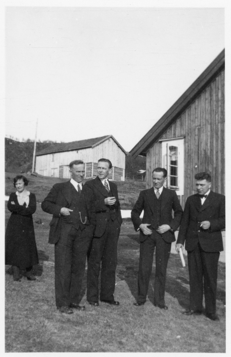 Kvinne og fire menn i penklær, fotografert foran gårdsbygninger.