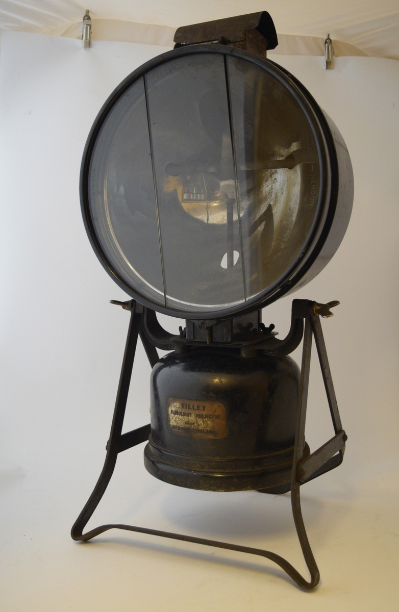 Lampe med parafinbeholder under selve lampen. Festet til stativ som gjør lampen stående og regulerbar i vinkel opp eller ned. Håndtak bak.