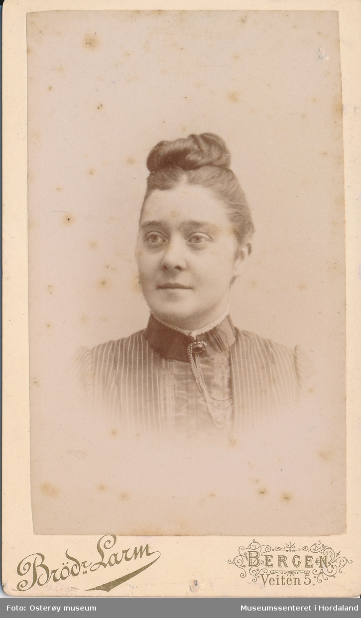 portrettfotografi av ung kvinne med håret i rull oppe på hove, plissert Kjole/bluse med solkekrage og brosje med kjede.