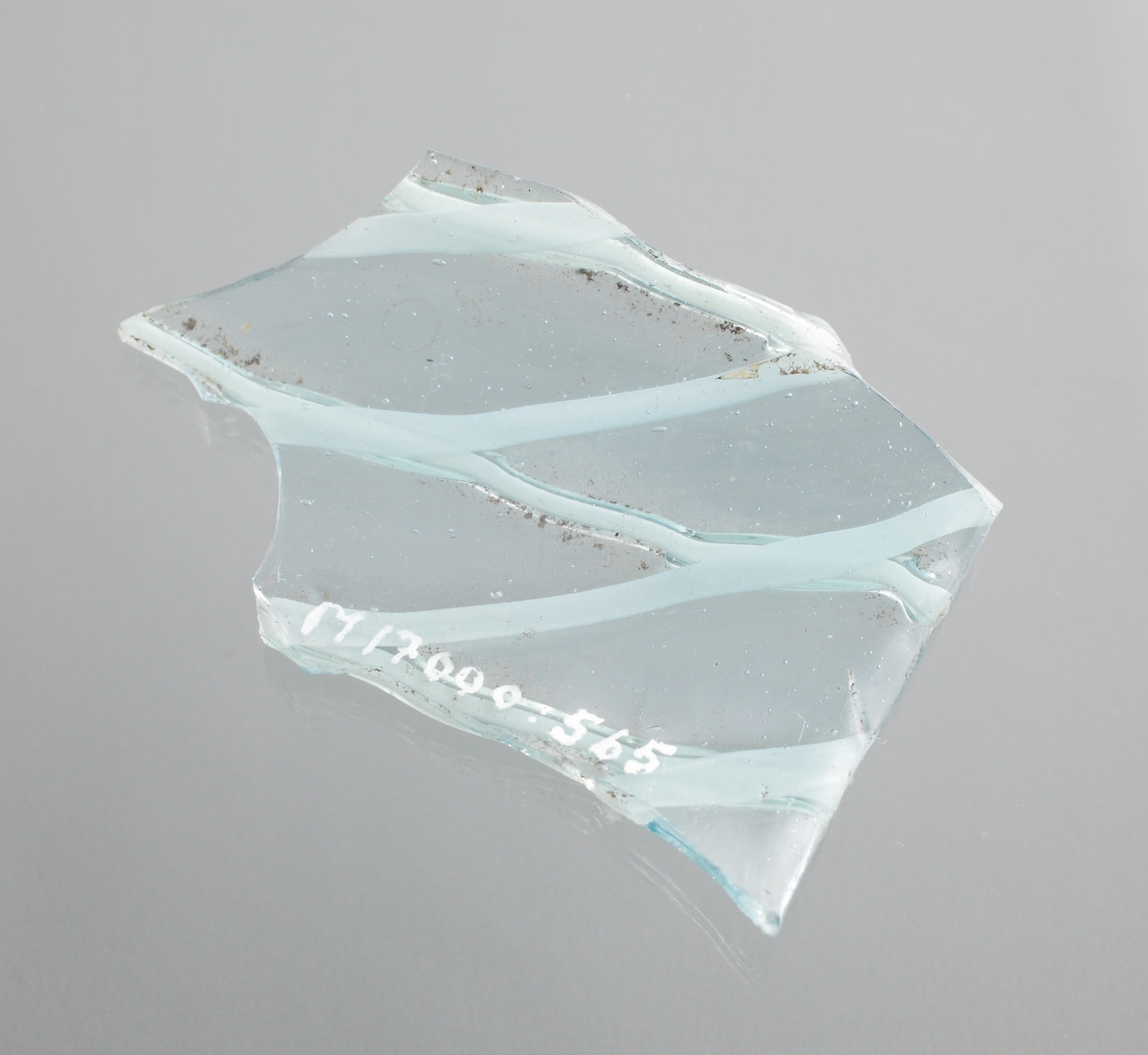 Två delar av ett ornerat venetianskt glas i svagt blå färg. På glaset finns två skikt pålagda vita trådar i ett grovt nätmönster (vetro a fili) i relief. Den större av skärvorna är en del av en mynningsrand i ett cylindriskt glas.
