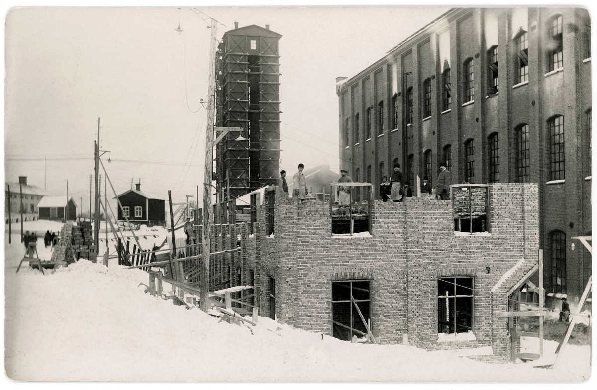 Uppförande av fabriksbyggnad vid Svartviks sulfitfabrik. Arbetslag som murar upp väggarna. I bakgrunden syratornet. Privattaget vykort.