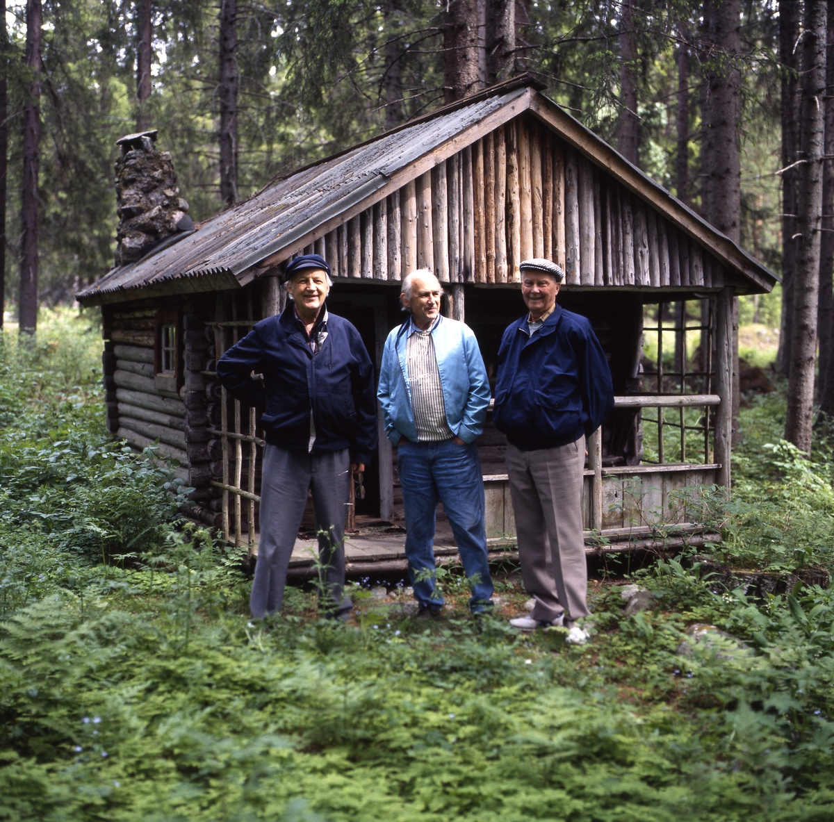 Några män, inklusive bröderna Ahnlund, står framför en gammal timmerstuga i en skogsglänta, 12 juni 1994.