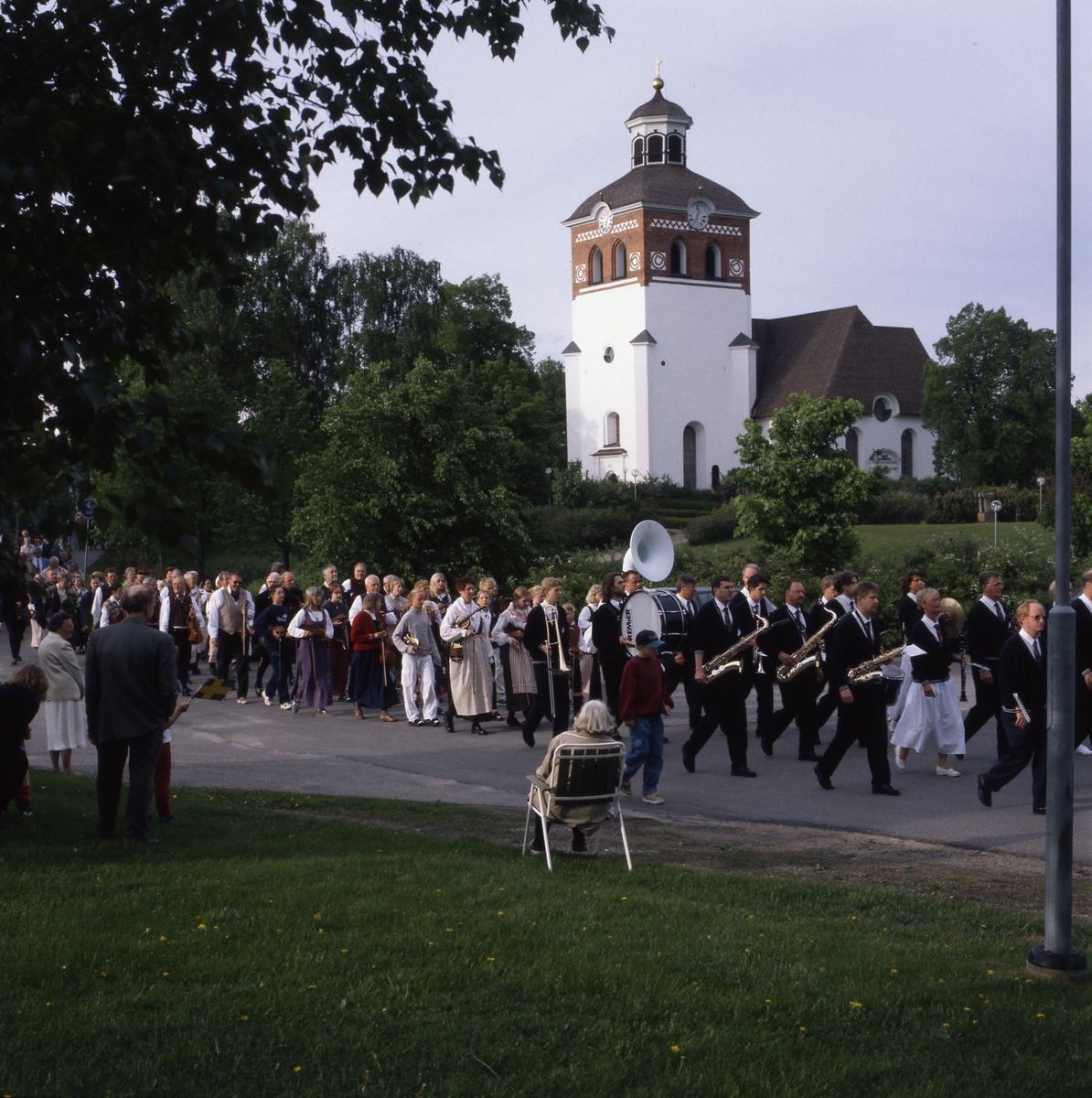Nationaldagen i Bollnäs 6 juni 1999. Fanbärare och musikanter tågar förbi kyrkan. En äldre kvinna sitter i en trädgårdsstol bredvid vägen och ser på.