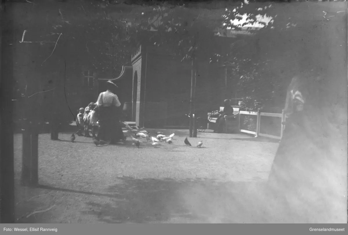 En kvinne og flere barn mater duer på en åpen plass, Oslo? En kvinne med barnevogn sitter på en benk og ser på.