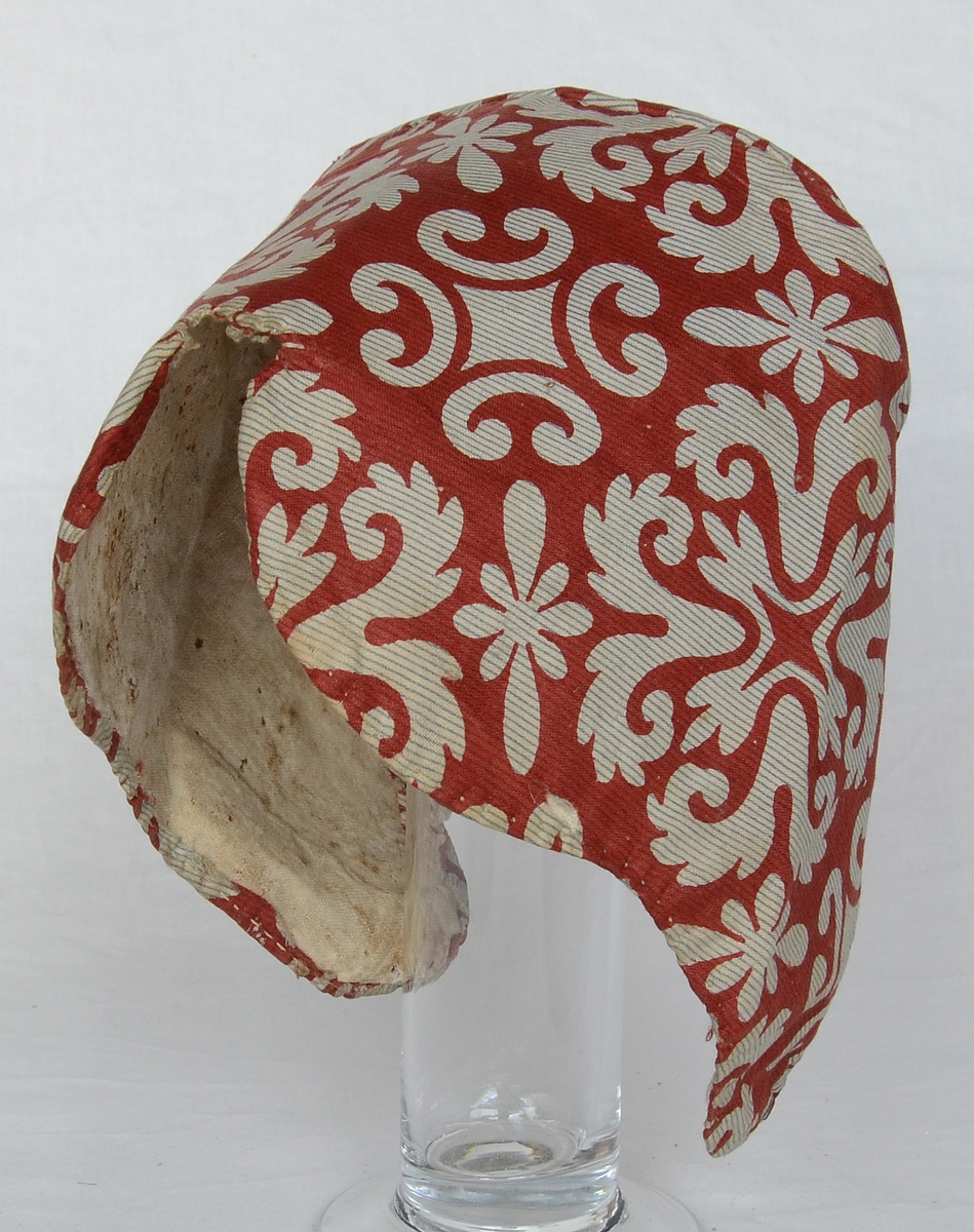 "Flatmössa" med linnefoder i tuskaftsbindning och mönstrat bomullstyg i rödbrunt, vitt och blått utanpå. Mössan är sydd med nackstycke.