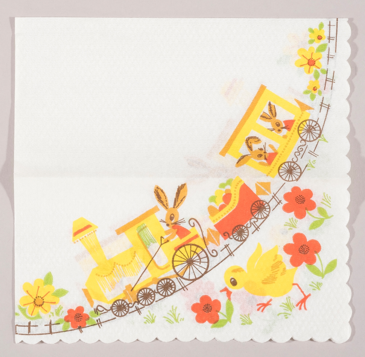 Et tog med lokomotiv og to vogner. En påskeharer sitter i lokomotivet og to i en vogn. I en tredje vogn er der påskeegg. En kylling løper i en blomstereng med røde og gule blomster.