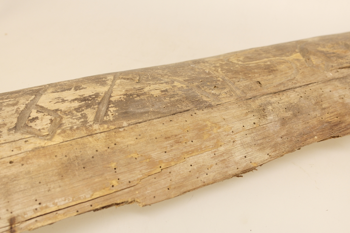 Kløyvet tømmerstokk i furu. I stokken er det skåret inn tall og bokstaver. På baksiden av stokken er det skrevet et årstall med blyant.