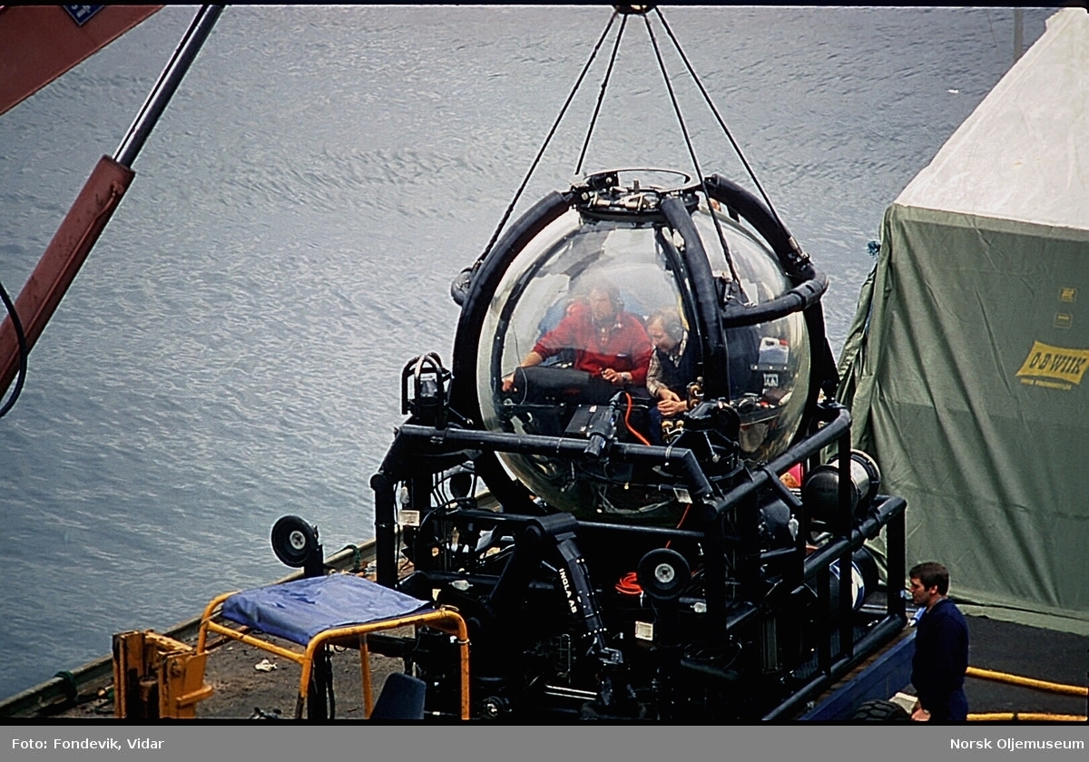To menn sitter i miniubåten Check Mate, utviklet av NUI/NUTEC.  Ubåten står på kaikanten ved NUI's base, og er festet til en kran.