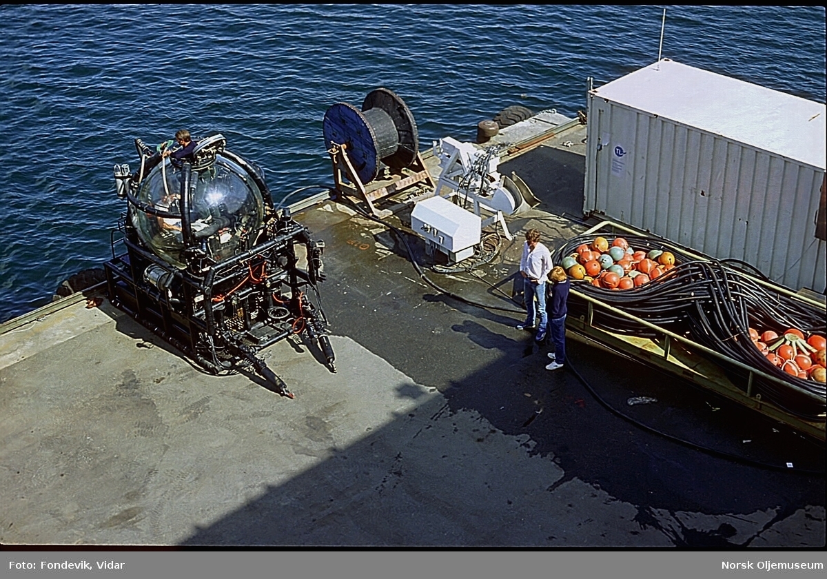 Miniubåten "Check Mate" og en heisekran står på kaien ved NUI i Gravdal.  I forgrunnen ligger en kabelbunt med påsatte blåser som brukes mellom ubåten og dykkekontrollen på moderskipet.
