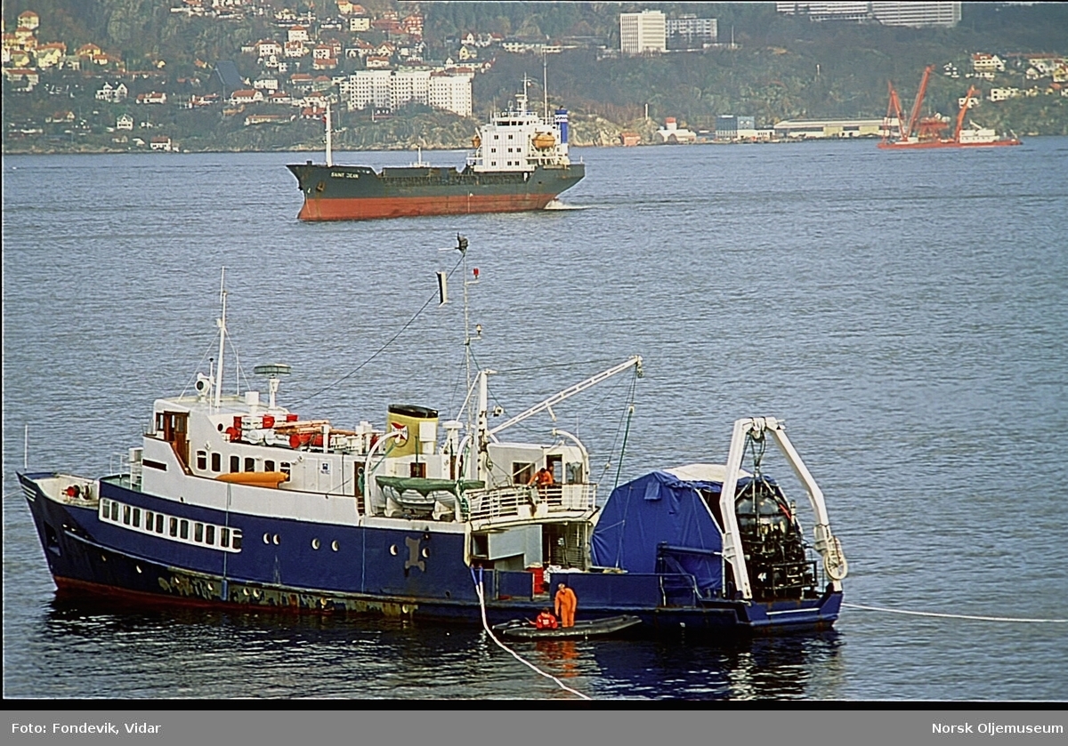 Dykkerskipet "Strilborg" ligger fortøyd i fjorden utenfor Bergen med miniubåten Check Mate hengende i akterenden.  Mannskap arbeider ombord.