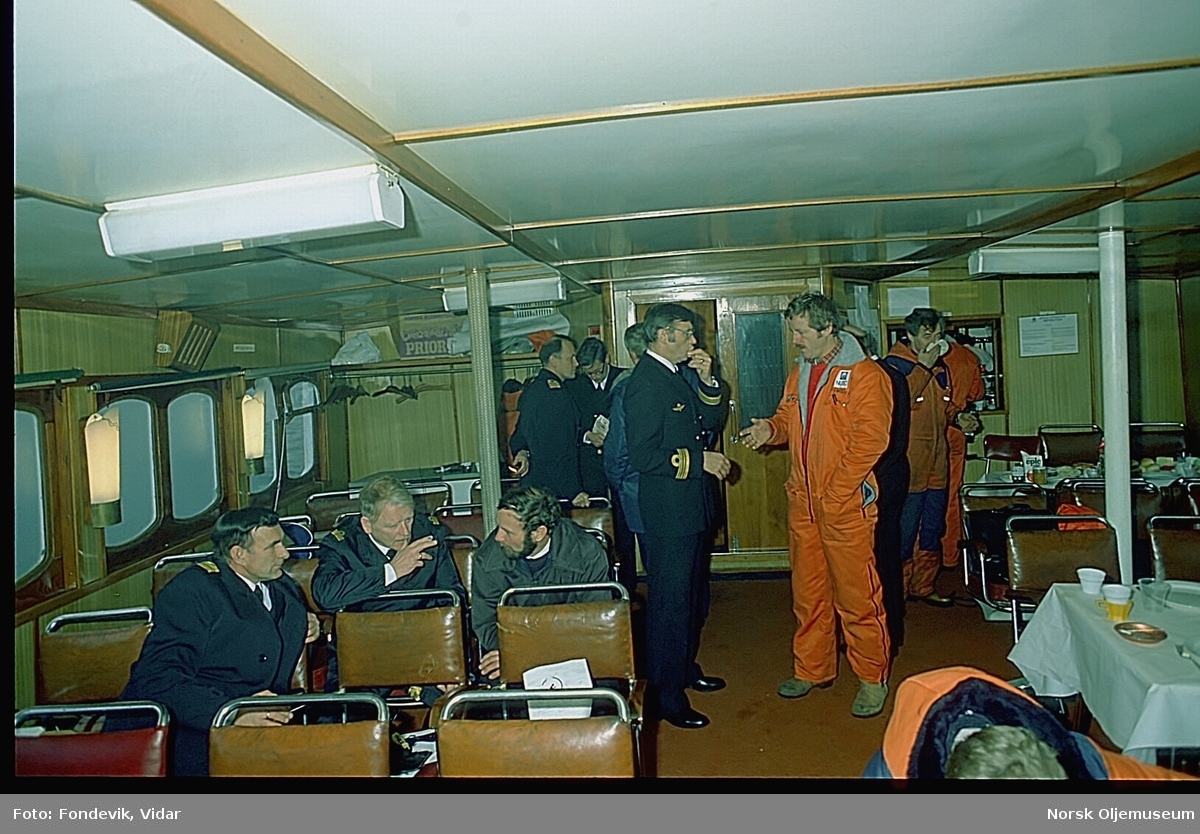 Bilder fra salongen til et skip. Det er flere menn samlet i salongen, hvorav flere av dem har uniformer fra Den kongelige norske marinen, og noen har arbeidsklær med logoen til "NUTEC" på.
Bildene kan muligens være tatt ombord fartøyet M/S "Halsnøy", bygd i 1957, for HSD