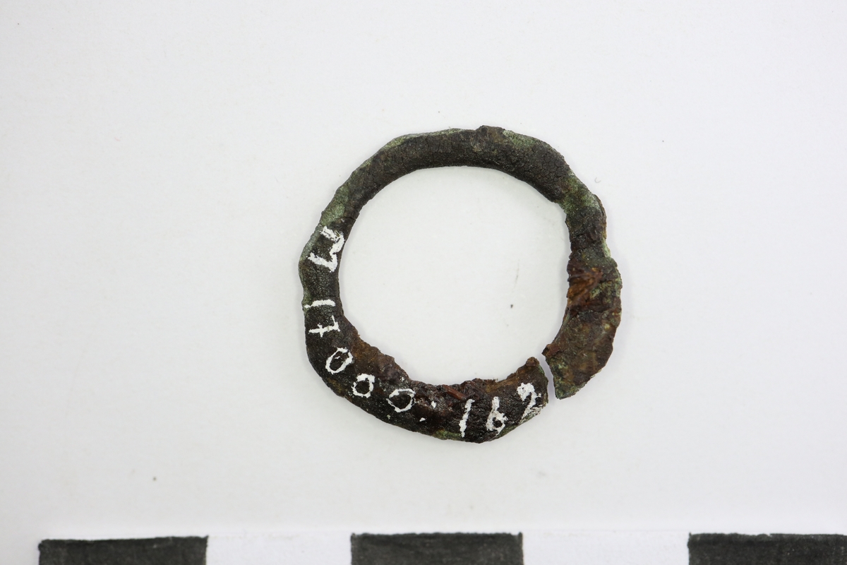 Fingerring av brons som är ornerad med graverade snedstreck (repslagsimitation?). Den övre delen av ringen är något högre än den undre. Förorenad med rost och små trärester.