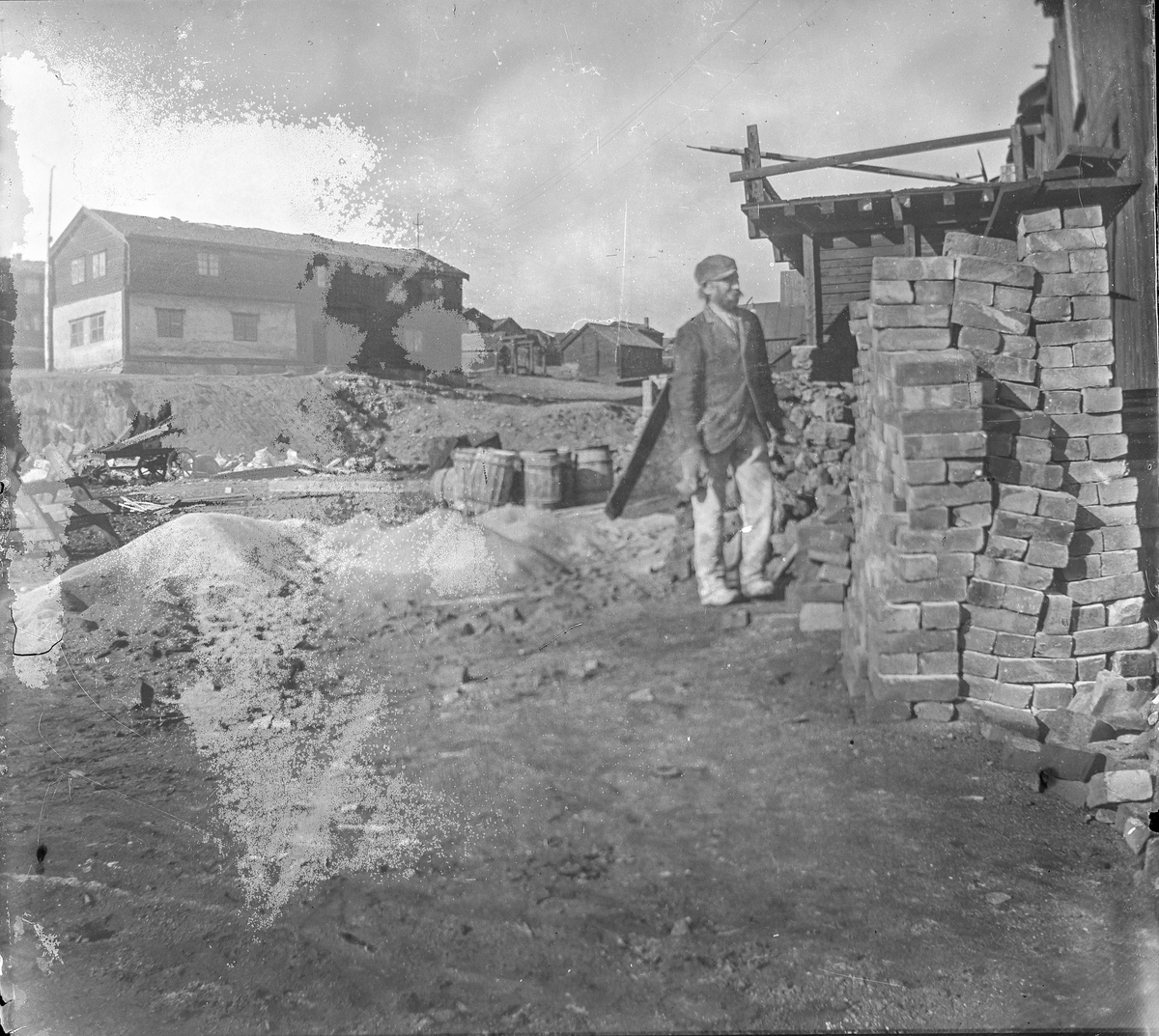 Fra Malmplassen, Røros. En arbeider står ved en stabel med murstein. Hyttstuggu i bakgrunnen