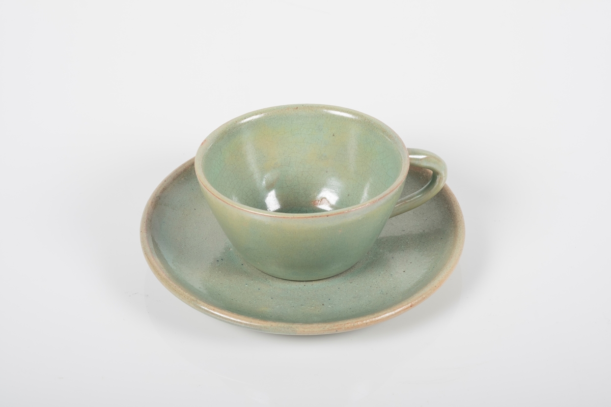 Kopp med skål i keramikk med grønn lasur. Buet hank på koppen. To små knotter (mangler en tredje knott) på undersiden av koppen, usikker funksjon. Skålen er blank på oversiden og matt på undersiden.
