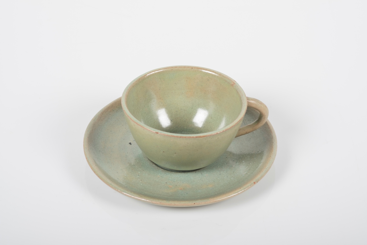 Kopp med skål i keramikk med grønn lasur. Buet hank på koppen. Koppen og skålen har spor etter tre knotter på bunnen, usikker funksjon. Bunnen på skålen har matt overflate.