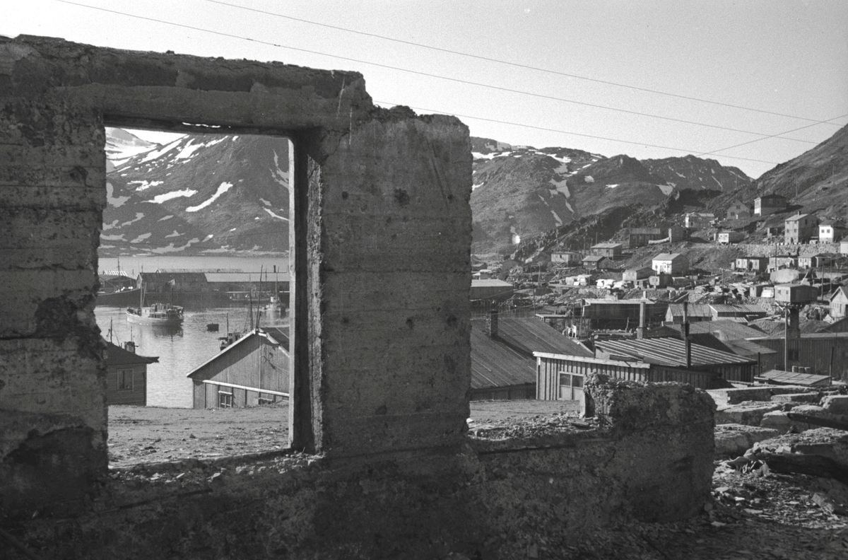 Gjenreisning. Deler av Honningsvåg med havn, brakkebebyggelsen og nybygde boliger, sett gjennom rester av et ødelagt hus. 1946/47.
