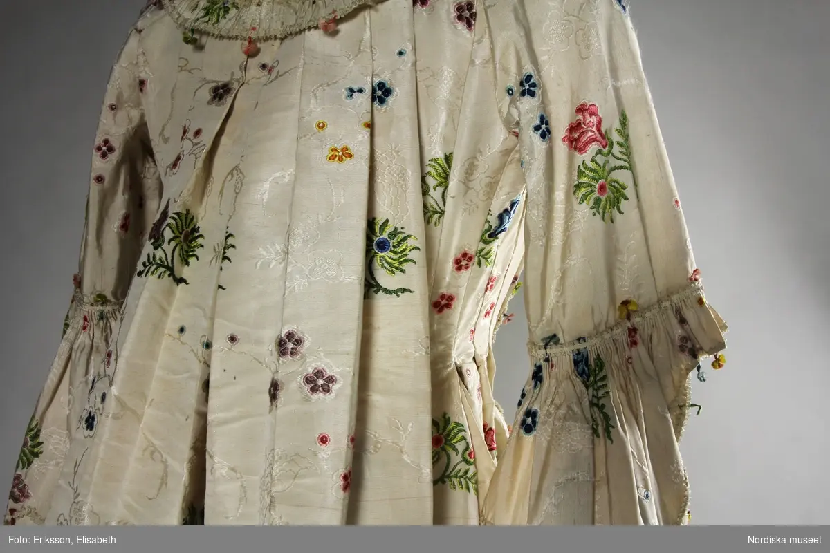 Brudklänning (robe), av broscherat, vitbottnat kinesiskt siden. Nygjord framvåd av fint vitt bomullstyg med kjolvolang nedtill av robens tyg. Två lösa garneringsbårder.